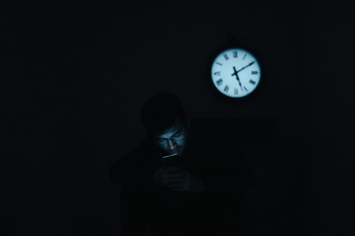 A person sitting still in dim light looking at their phone while a blurred clock on the wall symbolizes time passing — representing stagnation and delay
